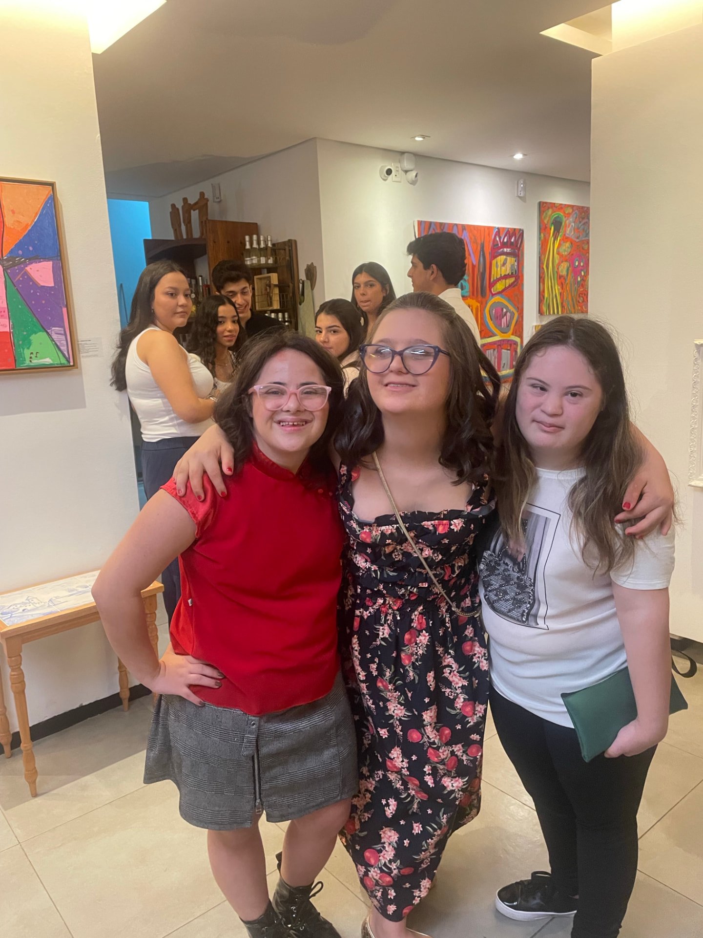 Três jovens abraçadas em uma exposição de arte, com quadros coloridos ao fundo.