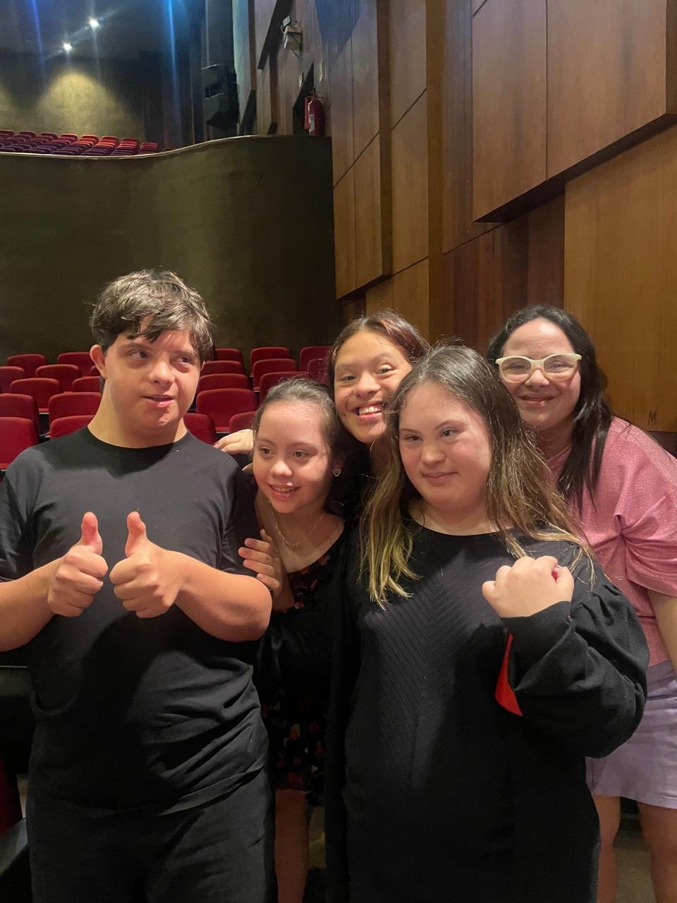 Grupo de jovens da APTIC no teatro, sorrindo e fazendo sinal de positivo depois da apresentação.